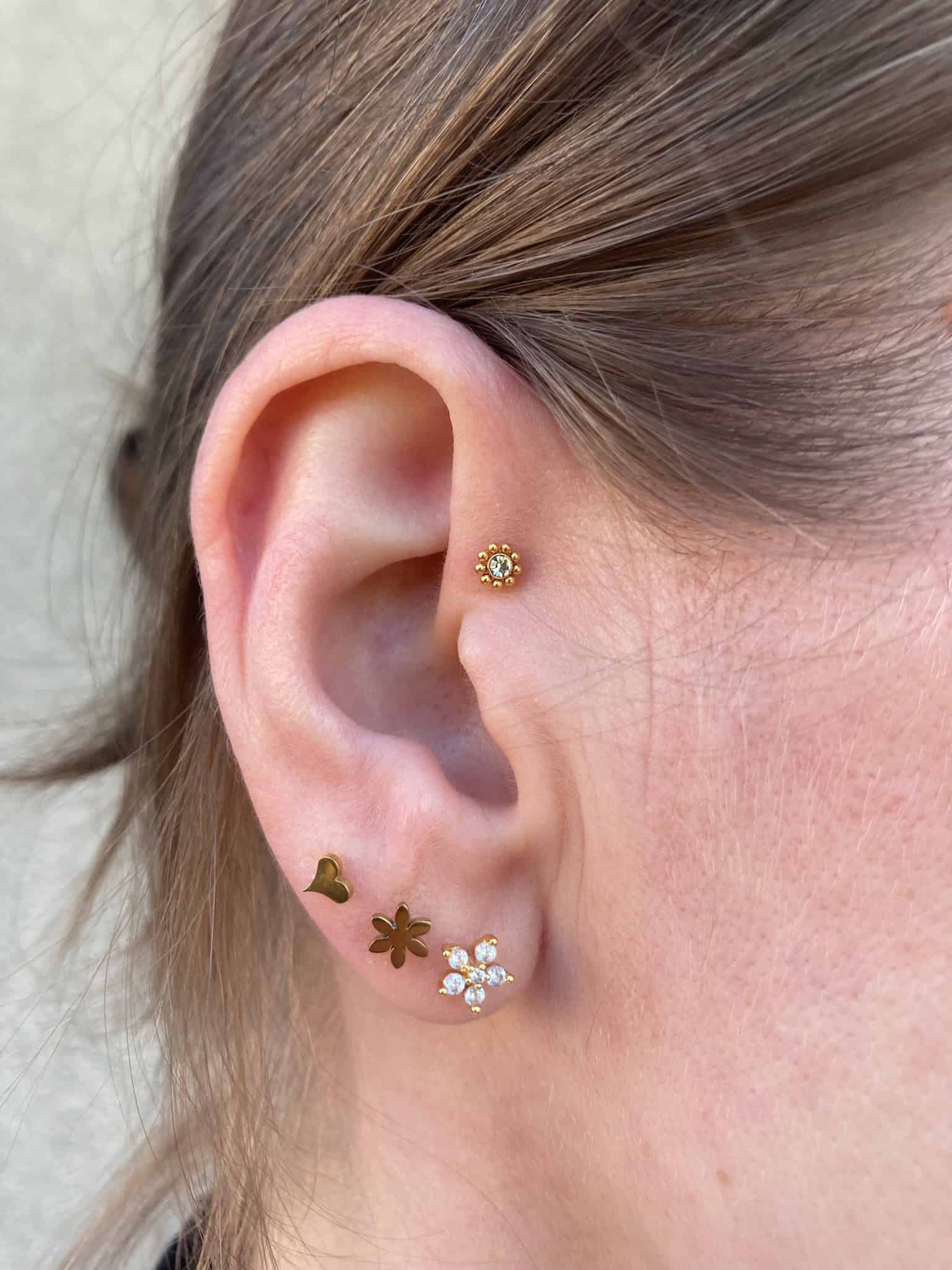 7sinstattoo.pl Zbliżenie ucha osoby ozdobionego wieloma piercingami, przedstawiające produkt IMG_6892. Górna chrząstka ucha prezentuje mały kolczyk w kształcie kwiatka, podczas gdy dolny płatek ucha prezentuje trzy kolczyki: małe serce, gwiazdę i kwiat ozdobiony kamieniami szlachetnymi. Brązowe włosy osoby są schowane za uchem, tworząc artystyczny wygląd przypominający studio tatuaży Poznań. Najlepsze studio tatuażu Seven Sins Tattoo