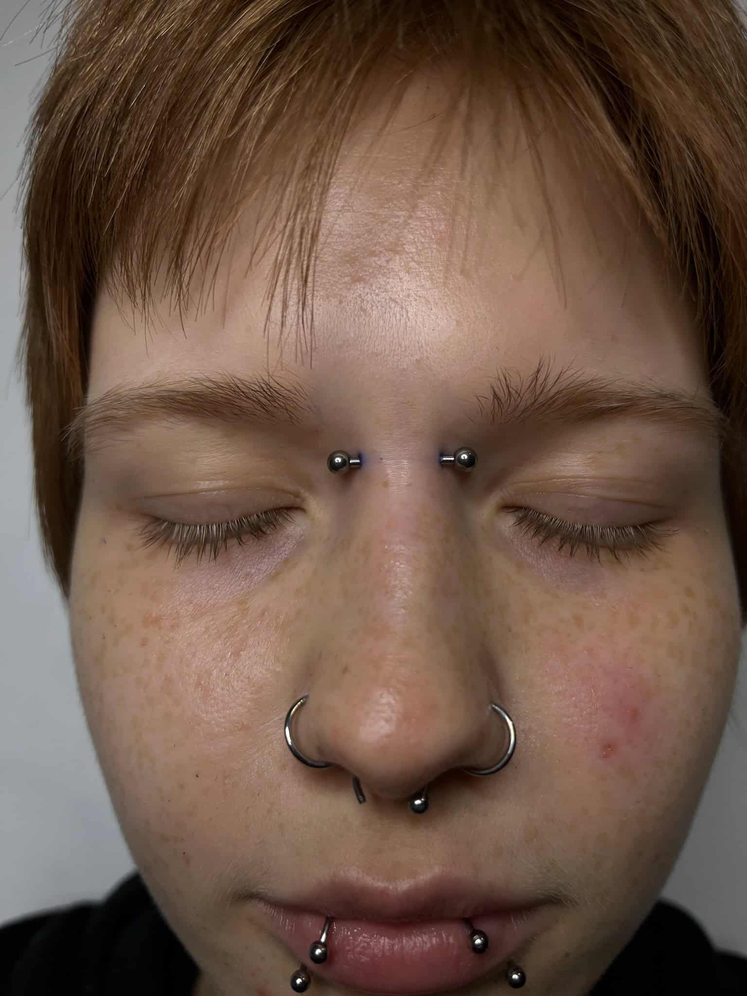 7sinstattoo.pl Osoba o krótkich włosach i jasnej karnacji zamyka oczy. Nosi wiele kolczyków na twarzy, w tym dwa nad grzbietem nosa, przekłucie przegrody nosowej, dwa na dolnej wardze i po jednym w każdym nozdrzu. Jej wyraz twarzy jest neutralny, prawdopodobnie rozważa swój kolejny projekt w studio tatuaży Poznań. Ta fotografia nosi tytuł IMG_6912. Najlepsze studio tatuażu Seven Sins Tattoo