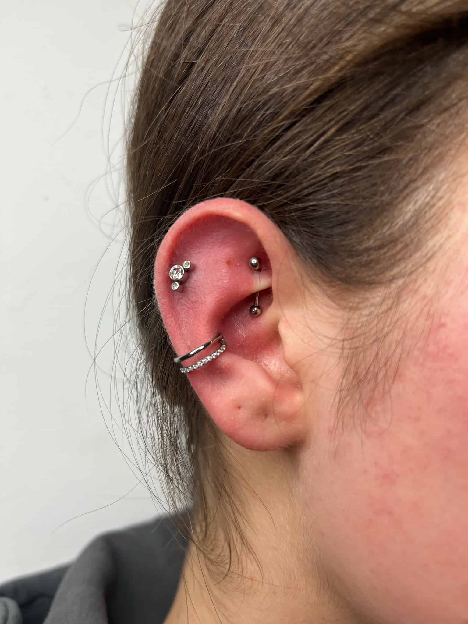 7sinstattoo.pl Zbliżenie ucha z wieloma piercingami ze studia tatuaży Poznań (IMG_7134). W chrząstce znajdują się dwa kolczyki-sztyfty, a w płatku dwa kolczyki-kółka, z których jeden ma rząd małych kamieni. Ucho ma delikatne zaczerwienienie wokół piercingu, widoczne są brązowe włosy i jasna skóra. Najlepsze studio tatuażu Seven Sins Tattoo