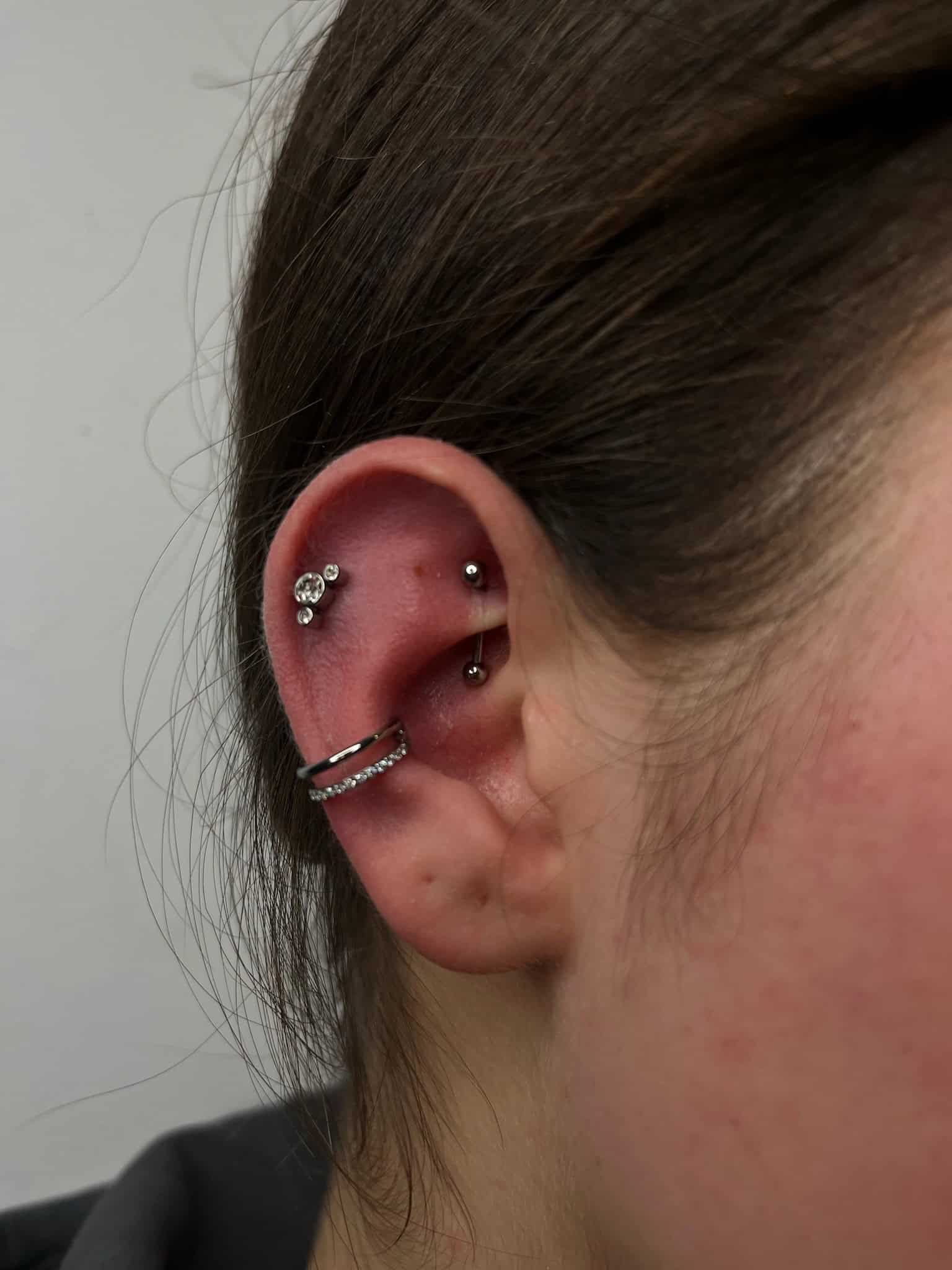 7sinstattoo.pl Zbliżenie na ucho osoby z wieloma piercingami, wykonane w najlepszym studio tatuażu w Poznaniu, z IMG_7136. Piercingi obejmują mały kolczyk z wzorem w kształcie kwiatu na górnej części ucha, zakrzywiony pręt w chrząstce i kolczyk-kółko z osadzonymi klejnotami w płatku ucha. Ucho jest osadzone na jednolitym tle. Najlepsze studio tatuażu Seven Sins Tattoo