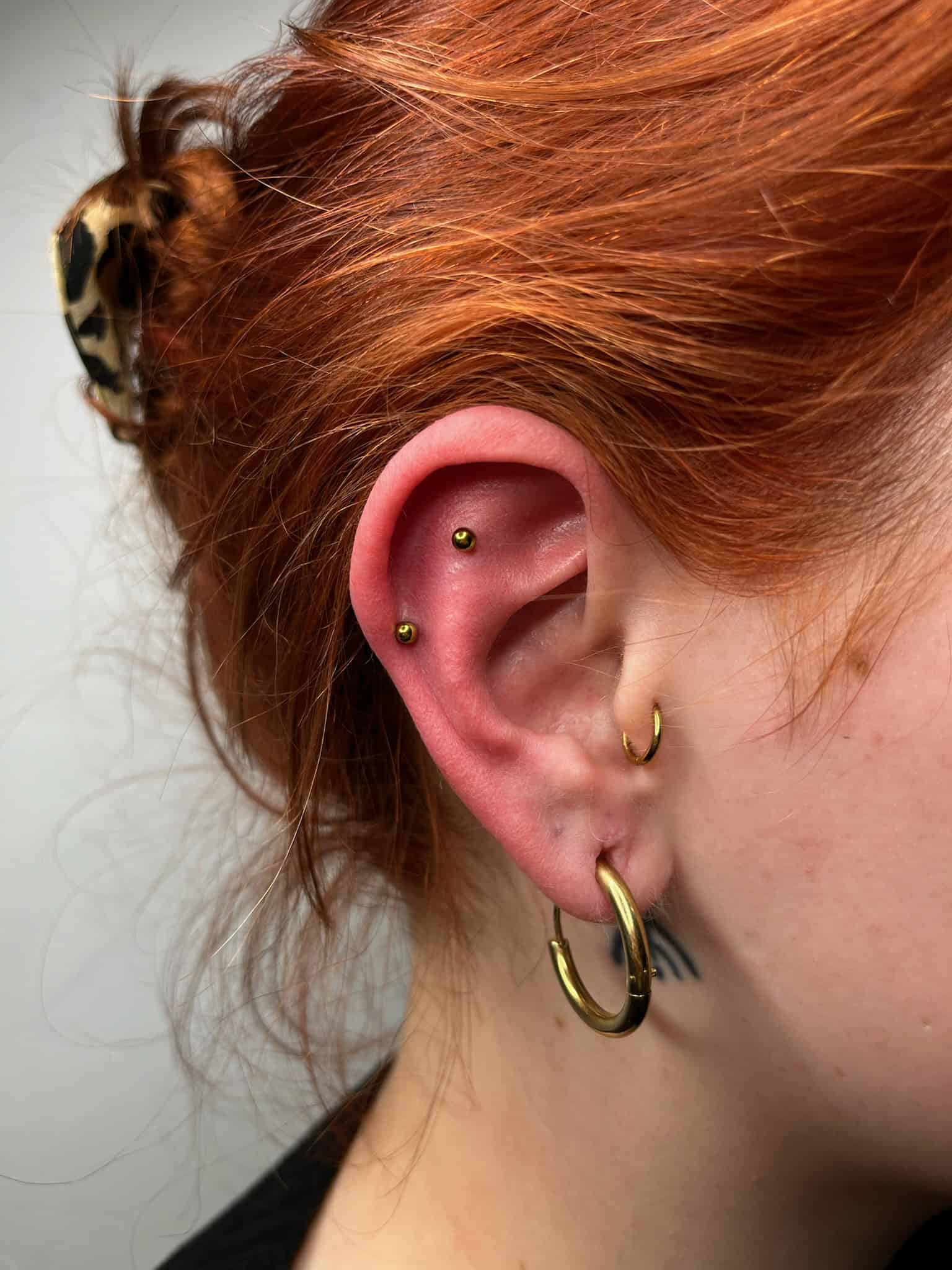 7sinstattoo.pl Zbliżenie ucha osoby z wieloma piercingami, prezentujące IMG_7293, w tym złoty kółko w płatku ucha, mniejszy kółko w górnym uchu i dwa złote kolczyki na wewnętrznej chrząstce. Osoba ma rude włosy związane brązową, wzorzystą gumką do włosów. Po inspirację do sztuki na ciele odwiedź Studio Tatuaże Poznań. Najlepsze studio tatuażu Seven Sins Tattoo