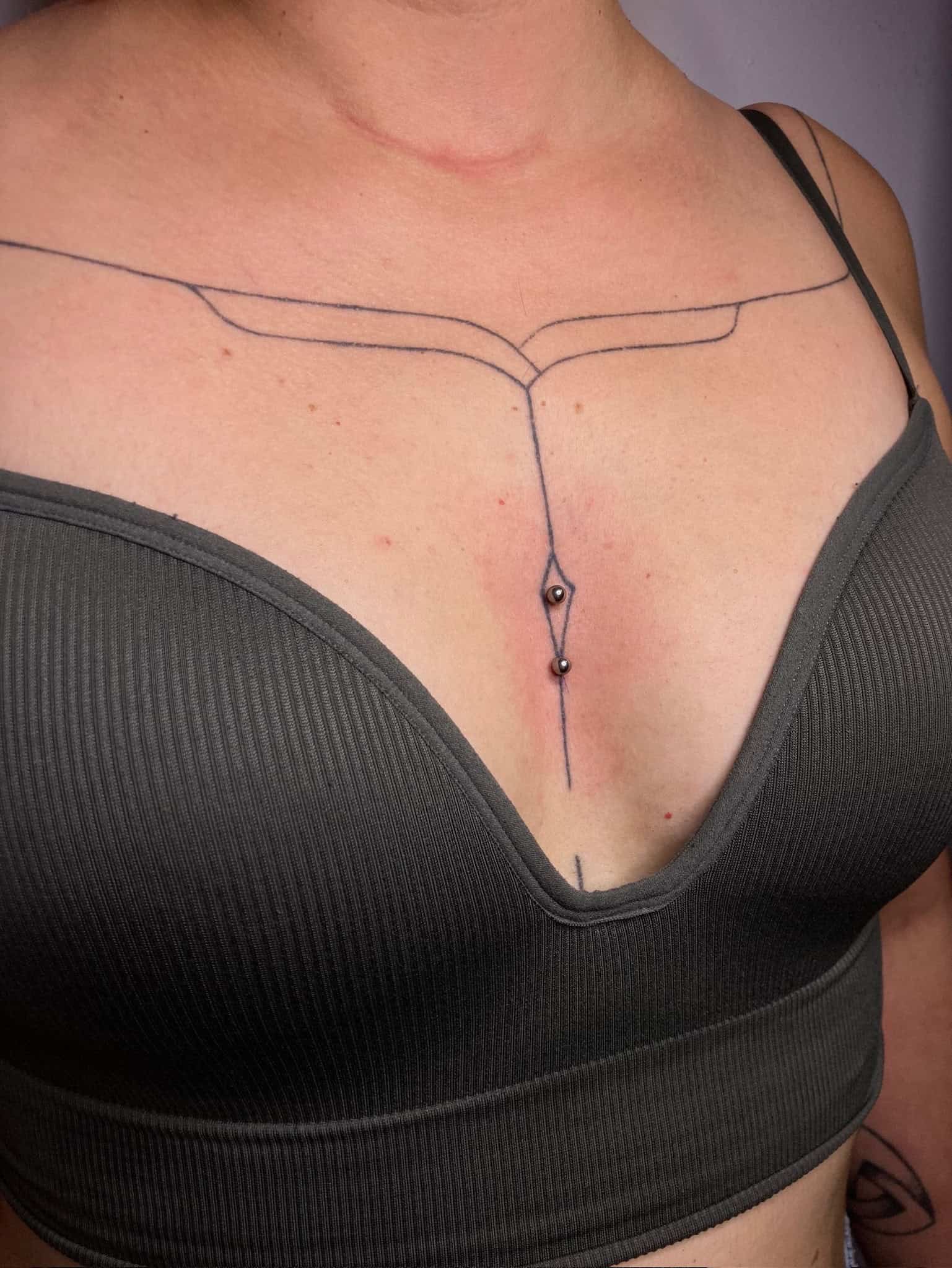 7sinstattoo.pl Osoba ubrana w IMG_7303, prążkowany, ciemnoszary podkoszulek, odsłania minimalistyczny tatuaż na klatce piersiowej ze studia tatuaży Poznań, przedstawiający linie rozciągające się przez obszar obojczyka i zbiegające się w spiczasty kształt w środku górnej części klatki piersiowej. Pod tatuażem widoczne są również dwa piercingi skórne. Najlepsze studio tatuażu Seven Sins Tattoo