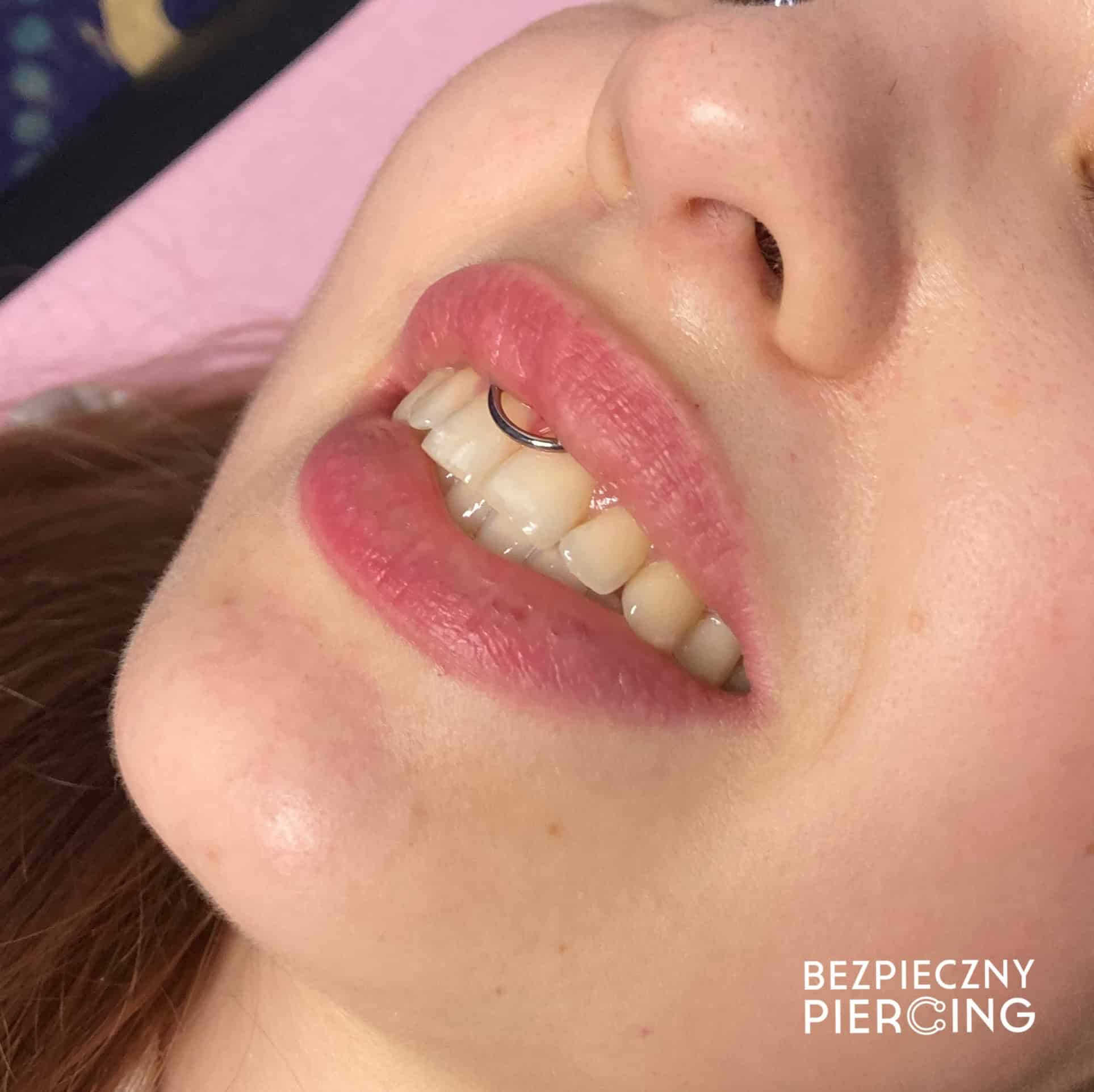7sinstattoo.pl Zbliżenie twarzy osoby pochylonej ku górze, ukazującej uśmiech ozdobiony srebrnym kółkiem w wardze i jasnoróżową szminką, z napisem „BEZPIECZNY PIERCING” w prawym dolnym rogu, ilustrującym wysokie standardy utrzymywane w studio tatuażu Poznań. Obraz nosi tytuł IMG_7516. Najlepsze studio tatuażu Seven Sins Tattoo