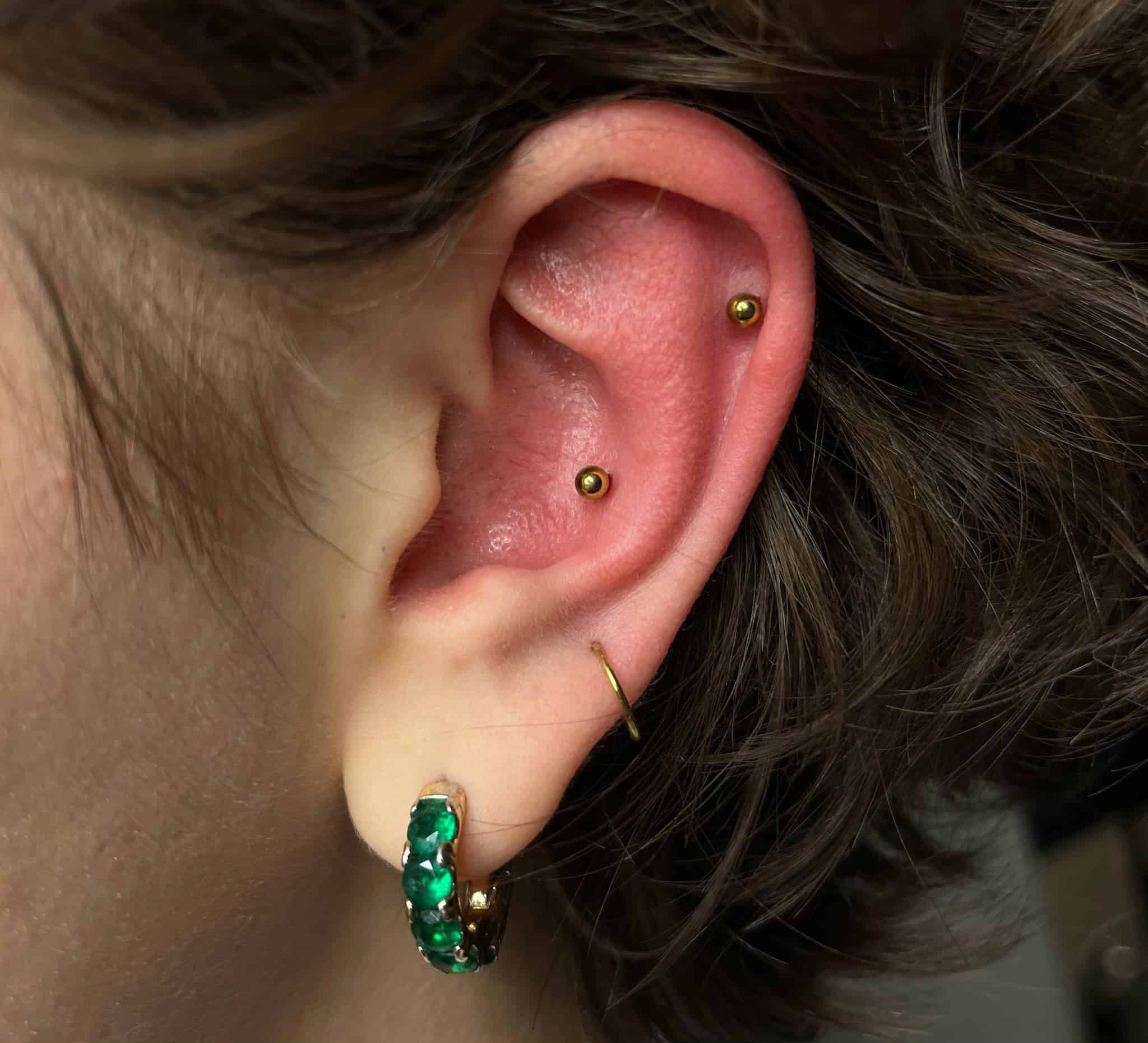 7sinstattoo.pl Zbliżenie ucha osoby ozdobionego wieloma piercingami, zrobione w jednym z najlepszych studiów tatuażu w Poznaniu. Ucho ma złoty kolczyk w okolicy muszli, złoty kolczyk w górnym płatku ucha, mały złoty pierścień w płatku ucha i zielony koralikowy kolczyk w kształcie kółka w dolnym płatku ucha. Obraz nosi tytuł IMG_8588. Najlepsze studio tatuażu Seven Sins Tattoo