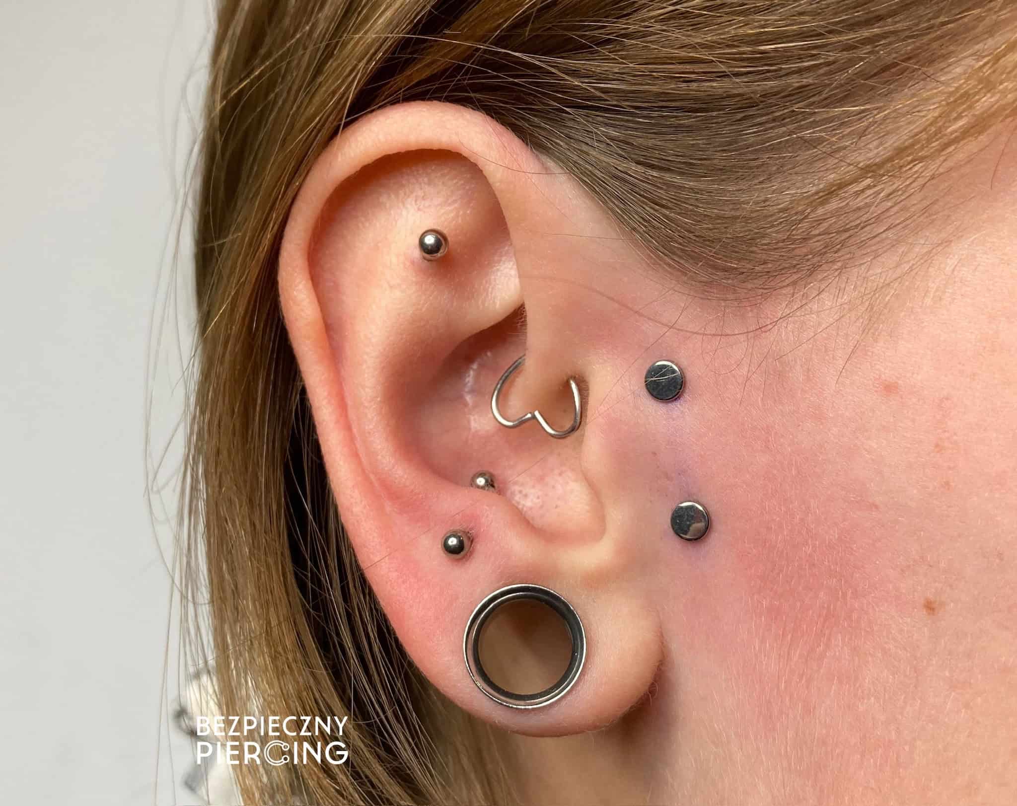 7sinstattoo.pl Zbliżenie ucha osoby z wieloma piercingami, w tym kółkiem w przedniej helisie, ćwiekiem w górnej helisie, kółkiem w muszli, ćwiekiem w tragusie i dwoma ćwiekami w płatku ucha wraz z tunelem. Tekst „BEZPIECZNY PIERCING” znajduje się w lewym dolnym rogu. Odwiedź nasze studio tat (Nazwa produktu: IMG_8634). Najlepsze studio tatuażu Seven Sins Tattoo