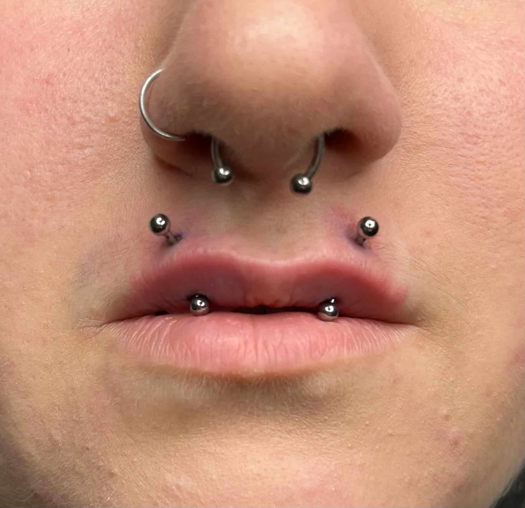 7sinstattoo.pl Zbliżenie ust i nosa osoby, prezentujące kilka piercingów. Osoba ma dwa piercingi po każdej stronie dolnej wargi i dwa w środku, a także piercing przegrody nosowej i kolczyk w nozdrzu po lewej stronie. Ten uderzający wygląd został dopracowany w studio tatuaży Poznań, jak uchwycono w produkcie IMG_8928. Najlepsze studio tatuażu Seven Sins Tattoo