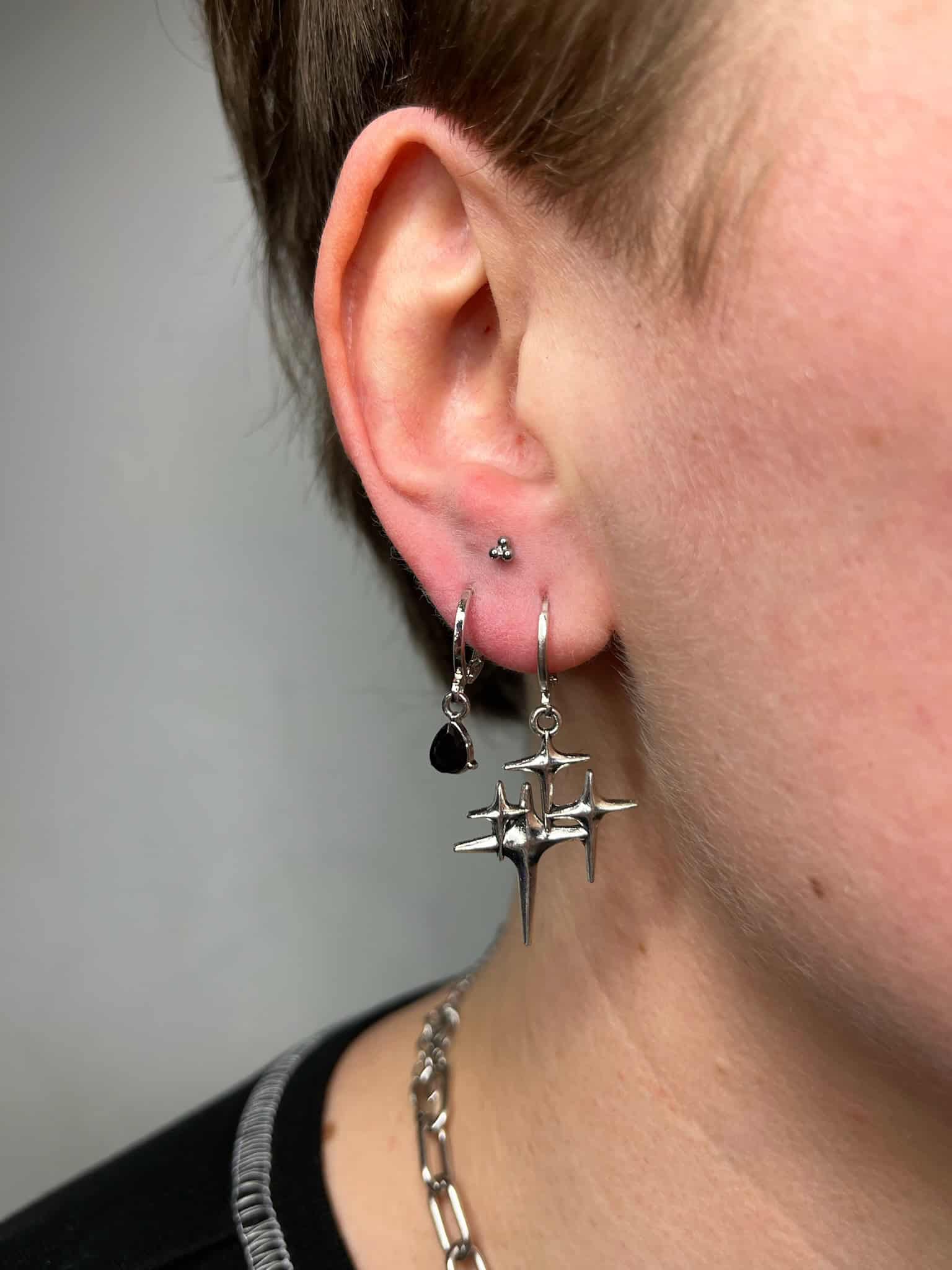 7sinstattoo.pl Zbliżenie ucha osoby ozdobionego dwoma kolczykami. Górny kolczyk to mały srebrny sztyft, podczas gdy dolny kolczyk prezentuje srebrny pierścień z czarnym łezkowatym urokiem i zwisającym geometrycznym srebrnym wzorem, jak pokazano na IMG_8987. Osoba, która często odwiedza najlepsze studio tatuażu Poznań, nosi również srebrny naszyjnik-łańcuszek. Najlepsze studio tatuażu Seven Sins Tattoo