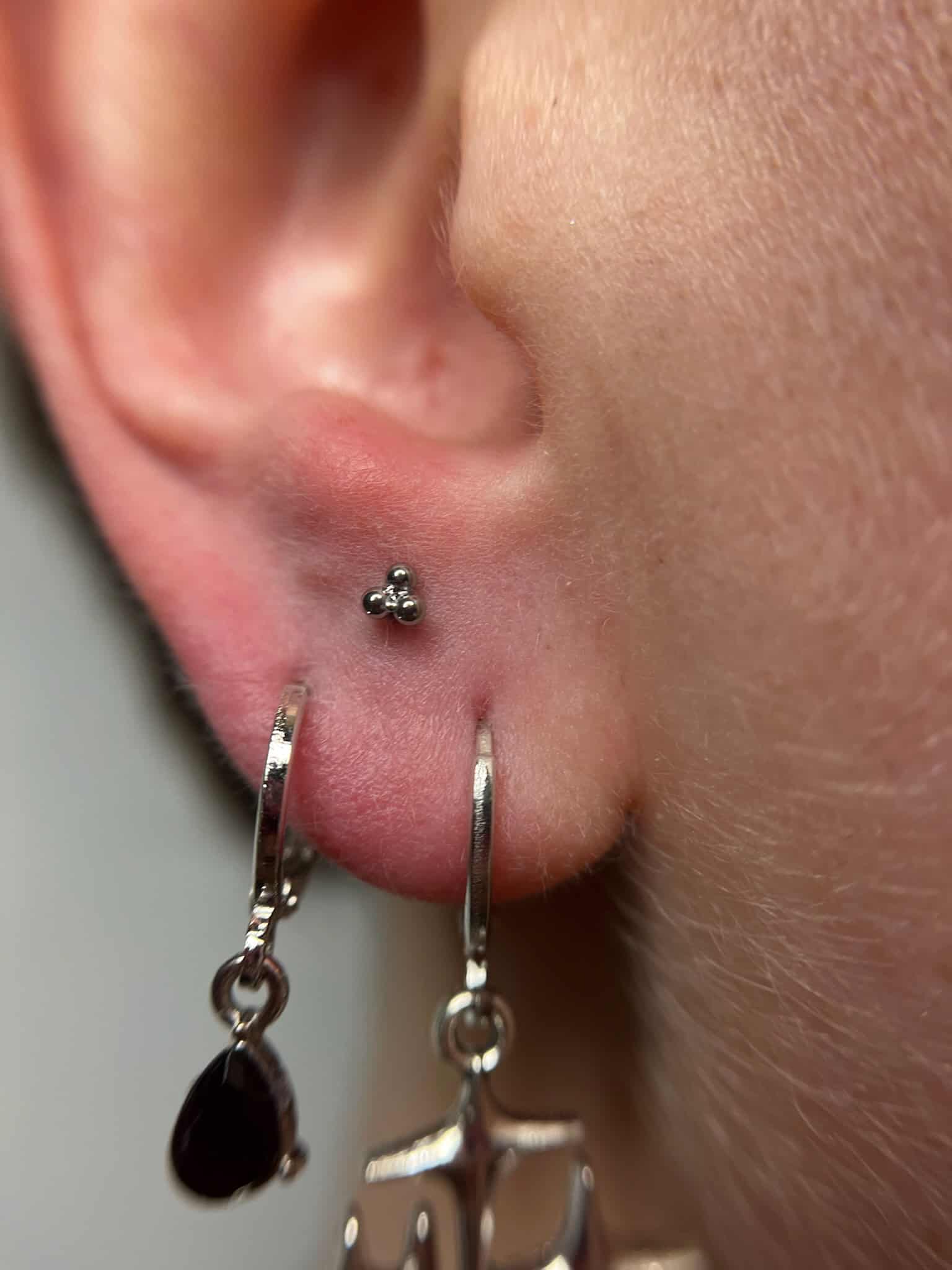 7sinstattoo.pl Zbliżenie na ucho z dwoma piercingami, ozdobione w studio tatuażu Poznań. Jeden piercing ma mały srebrny ćwiek z trzema malutkimi kulkami, podczas gdy drugi prezentuje IMG_9004, srebrny kolczyk-kółko z czarnym łezkowatym urokiem i zwisającym urokiem wagi. Najlepsze studio tatuażu Seven Sins Tattoo