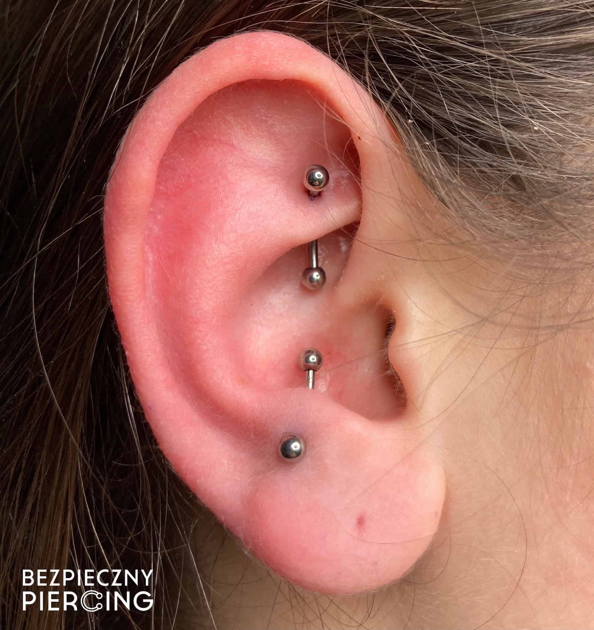 7sinstattoo.pl Zbliżenie na ucho osoby z wieloma piercingami, w tym trzema srebrnymi ćwiekami w chrząstce i jednym w płatku ucha. Tekst w lewym dolnym rogu brzmi „BEZPIECZNY PIERCING”. Skóra wokół piercingu wygląda na zdrową i dobrze zagojoną, co świadczy o doświadczeniu najlepszego studia tatuażu w Poznaniu. Obraz oznaczony jako IMG_9083. Najlepsze studio tatuażu Seven Sins Tattoo