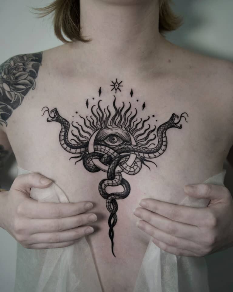 7sinstattoo.pl Osoba z dużym tatuażem na klatce piersiowej przedstawiającym oko otoczone płomieniami i splecionymi wężami trzyma lekką, półprzezroczystą tkaninę, częściowo zakrywającą dolną część tatuażu. Czarno-szary tatuaż kwiatowy, stworzony przez studio tatuażu Poznań, zdobi jej górną część lewego ramienia. Ten element nosi nazwę jsnhj. Najlepsze studio tatuażu Seven Sins Tattoo