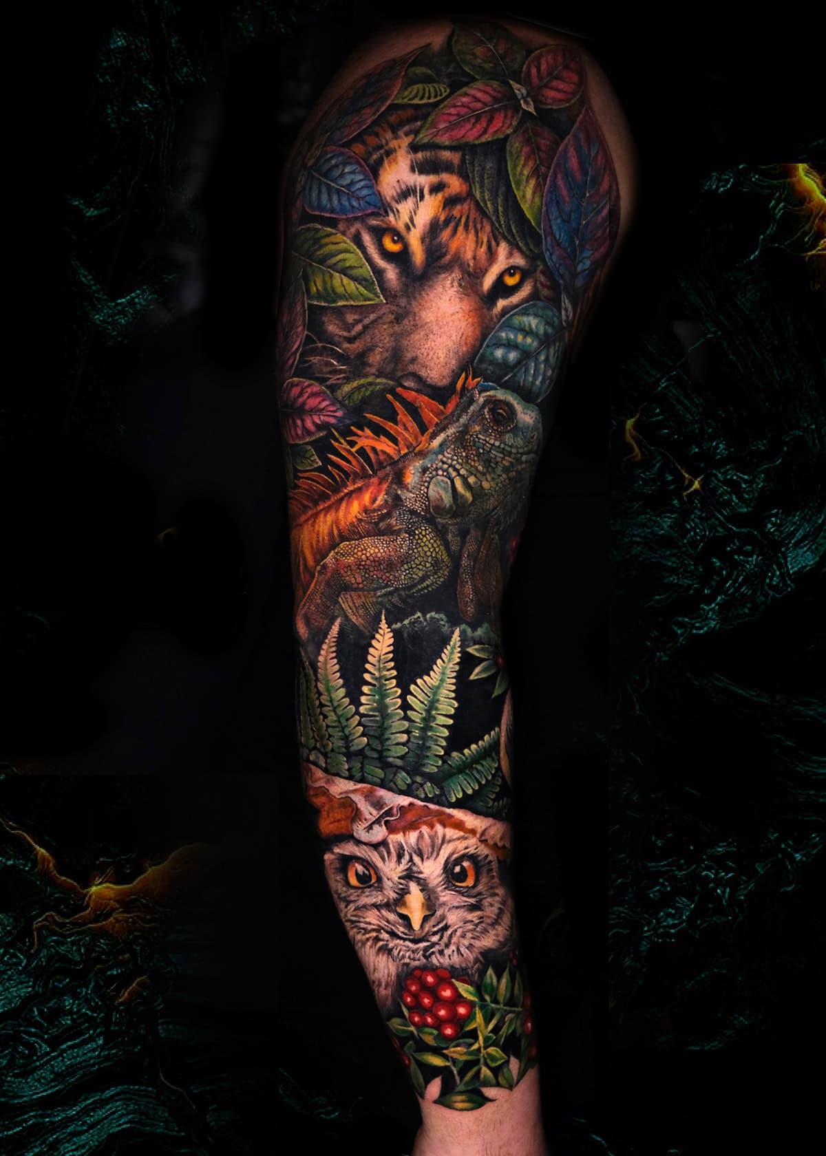 7sinstattoo.pl Żywy tatuaż pokrywający ramię, zatytułowany „IMG_7097” i stworzony przez utalentowanego tatuażystę z Poznania, przedstawia twarz tygrysa wśród liści u góry, iguanę na gałęzi pośrodku i sowę otoczoną liśćmi i jagodami u dołu. Ciemne tło podkreśla kolorowy wzór. Najlepsze studio tatuażu Seven Sins Tattoo