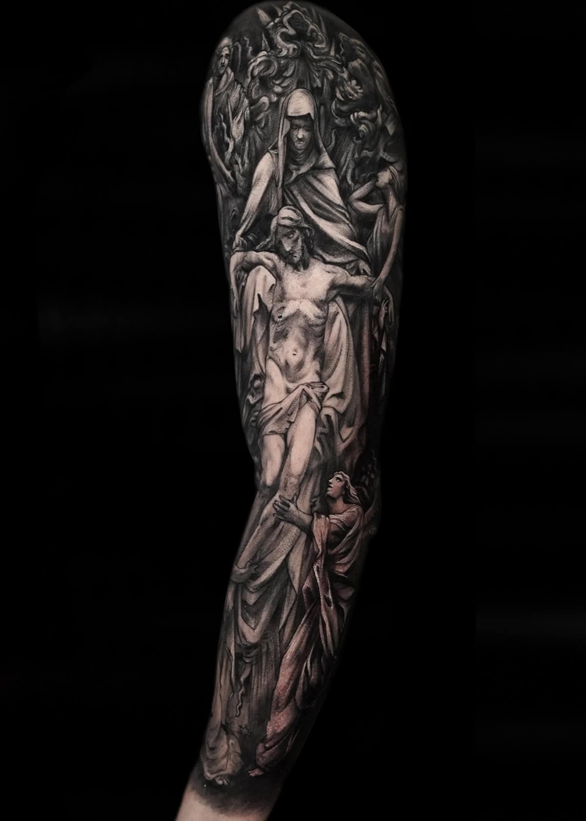 Szczegółowy czarno-biały tatuaż na rękawie przedstawia scenę religijną, z centralną postacią przypominającą Jezusa na krzyżu. Otaczające postacie to żałobnicy, prawdopodobnie Maryja, i różne anioły lub ikony religijne. Skomplikowany wzór pokrywa całe przedramię, rozciągając się do ramienia.