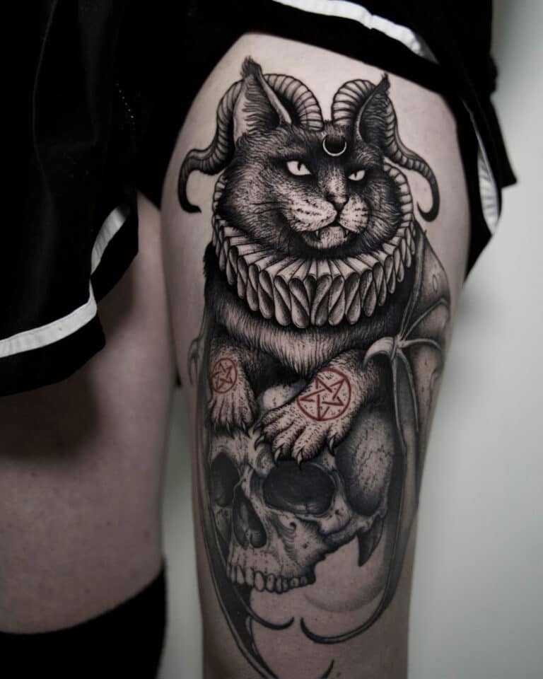 7sinstattoo.pl Szczegółowy tatuaż, uchwycony na zdjęciu produktu „Копия DSC09932” przez utalentowanego tatuażystę z Poznania, przedstawia kota z rogami barana i misterną obrożą, spoczywającego na ludzkiej czaszce. Na łapach kota znajdują się dekoracyjne symbole. Tatuaż jest umieszczony na udzie osoby, z widocznym brzegiem jej czarnego ubrania. Najlepsze studio tatuażu Seven Sins Tattoo