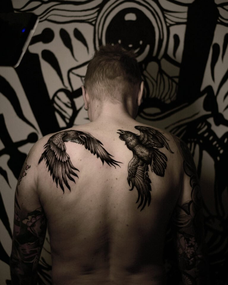 7sinstattoo.pl Osoba z krótkimi włosami i widocznymi tatuażami na ramionach jest widziana od tyłu, prezentując dwa duże tatuaże ptaków na górnej części pleców. Tło przedstawia czarno-białą abstrakcyjną sztukę muralową. Obraz ma słabe, dramatyczne oświetlenie, przedstawiające wykwintną pracę najlepszego studia tatuażu Poznań. Najlepsze studio tatuażu Seven Sins Tattoo