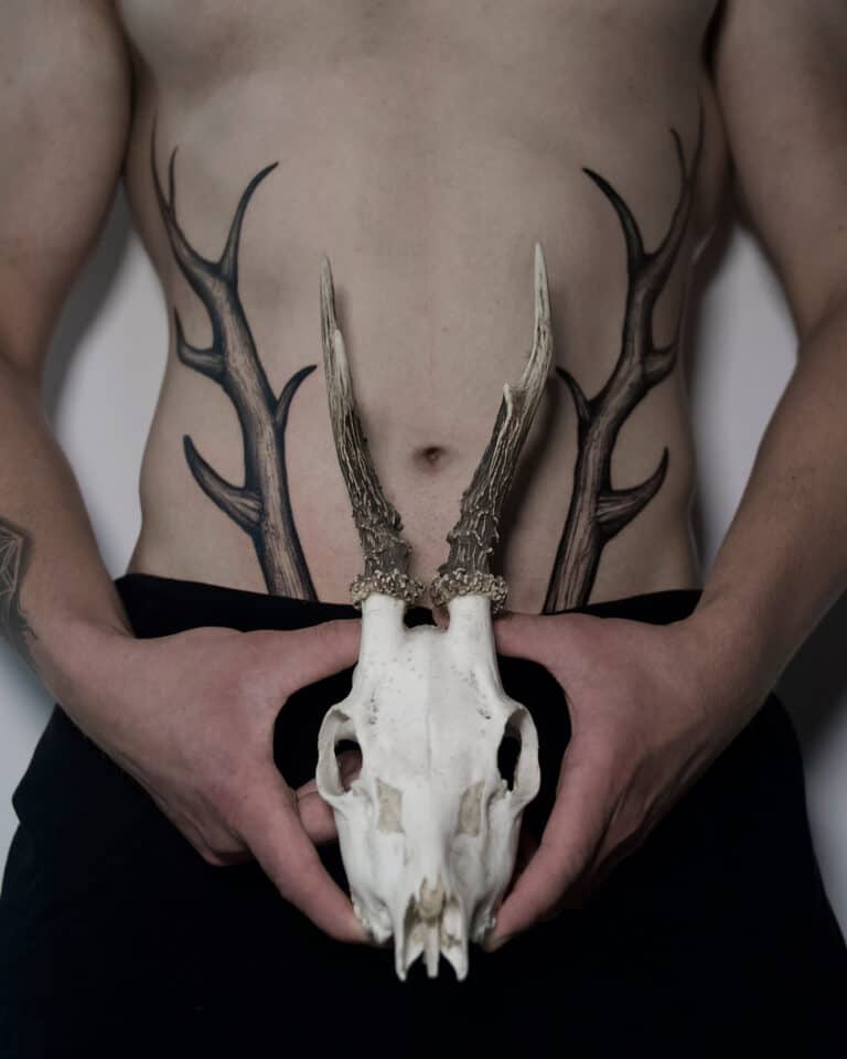 7sinstattoo.pl Osoba bez koszulki trzyma w rękach czaszkę jelenia, wyrównując jej poroże z misternymi czarnymi tatuażami poroża na brzuchu, tworząc iluzję, w której tatuaże płynnie rozciągają się od czaszki. Mają na sobie ciemne spodnie i stoją w renomowanym studiu tatuażu w Poznaniu. Ta artystyczna scena pięknie uosabia unikalny styl прррр. Najlepsze studio tatuażu Seven Sins Tattoo