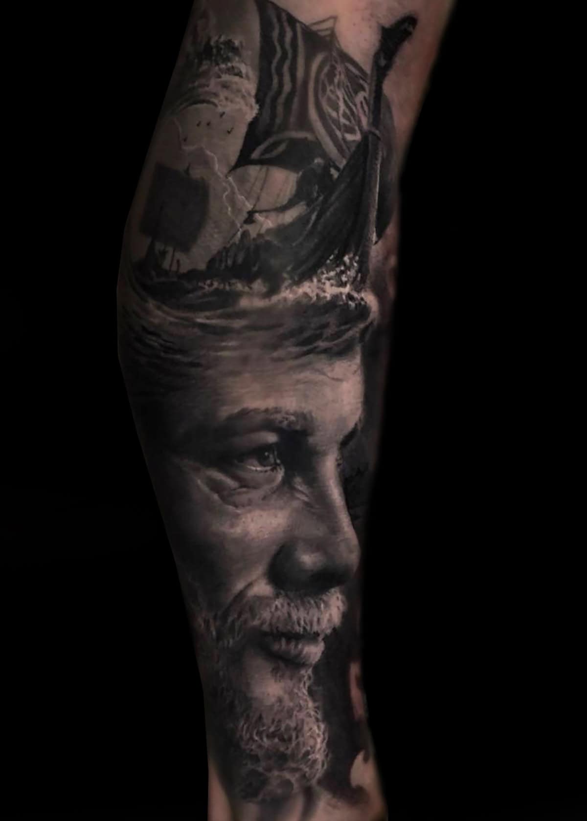 Tatuaż przedstawia szczegółowy portret w skali szarości starszego mężczyzny z brodą i głębokim spojrzeniem na przedramieniu. Nad mężczyzną znajduje się przedstawienie statku wikingów z rozwiniętymi żaglami, dodając element historyczny. Cały tatuaż ostro kontrastuje z czarnym tłem.