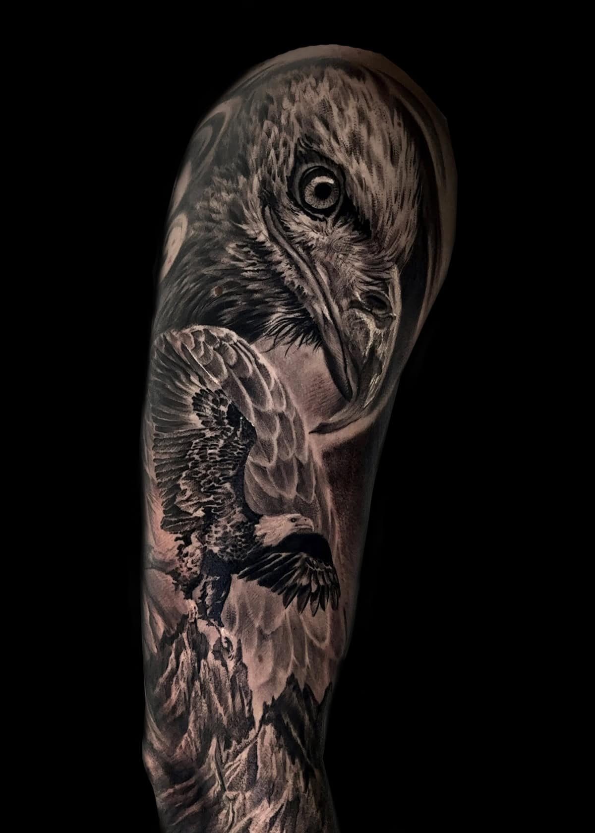 Szczegółowy czarno-szary tatuaż rozciąga się na górnej części ramienia, przedstawiając realistyczną głowę orła i drugiego orła w locie nad górzystym krajobrazem. Skomplikowany projekt charakteryzuje się realistycznym cieniowaniem i fakturami, podkreślającymi pióra i dramatyczne tło.