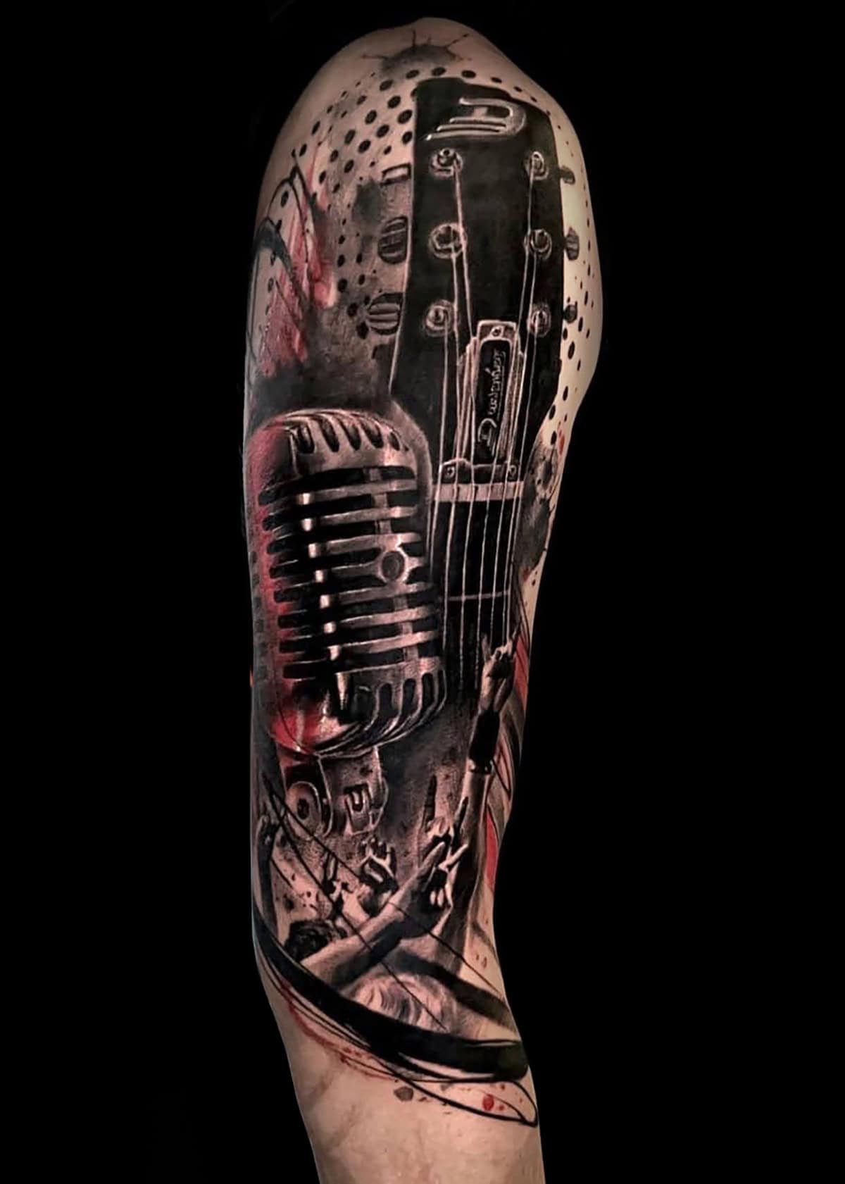 Szczegółowy czarno-szary tatuaż na ramieniu przedstawia retro mikrofon i część gitary. Artystyczne plamy czerwieni i bieli są zintegrowane z projektem, dodając żywego kontrastu i głębi. Grafika umiejętnie łączy różne tekstury, dając dynamiczny i dramatyczny efekt wizualny.