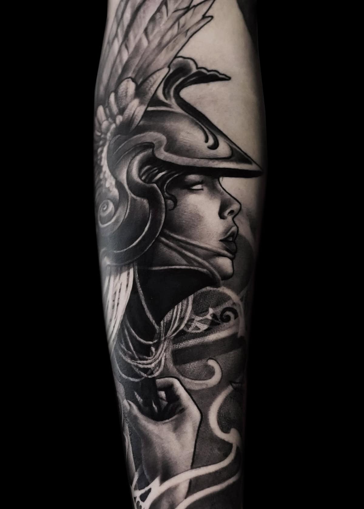 Czarno-szary tatuaż przedstawia misternie szczegółową wojowniczkę w skrzydlatym hełmie ozdobionym piórami, patrzącą w prawo z pogodnym wyrazem twarzy. Zbroja wojowniczki obejmuje eleganckie wzory, a płynne linie sugerują ruch lub wiatr, tworząc dynamiczną kompozycję.