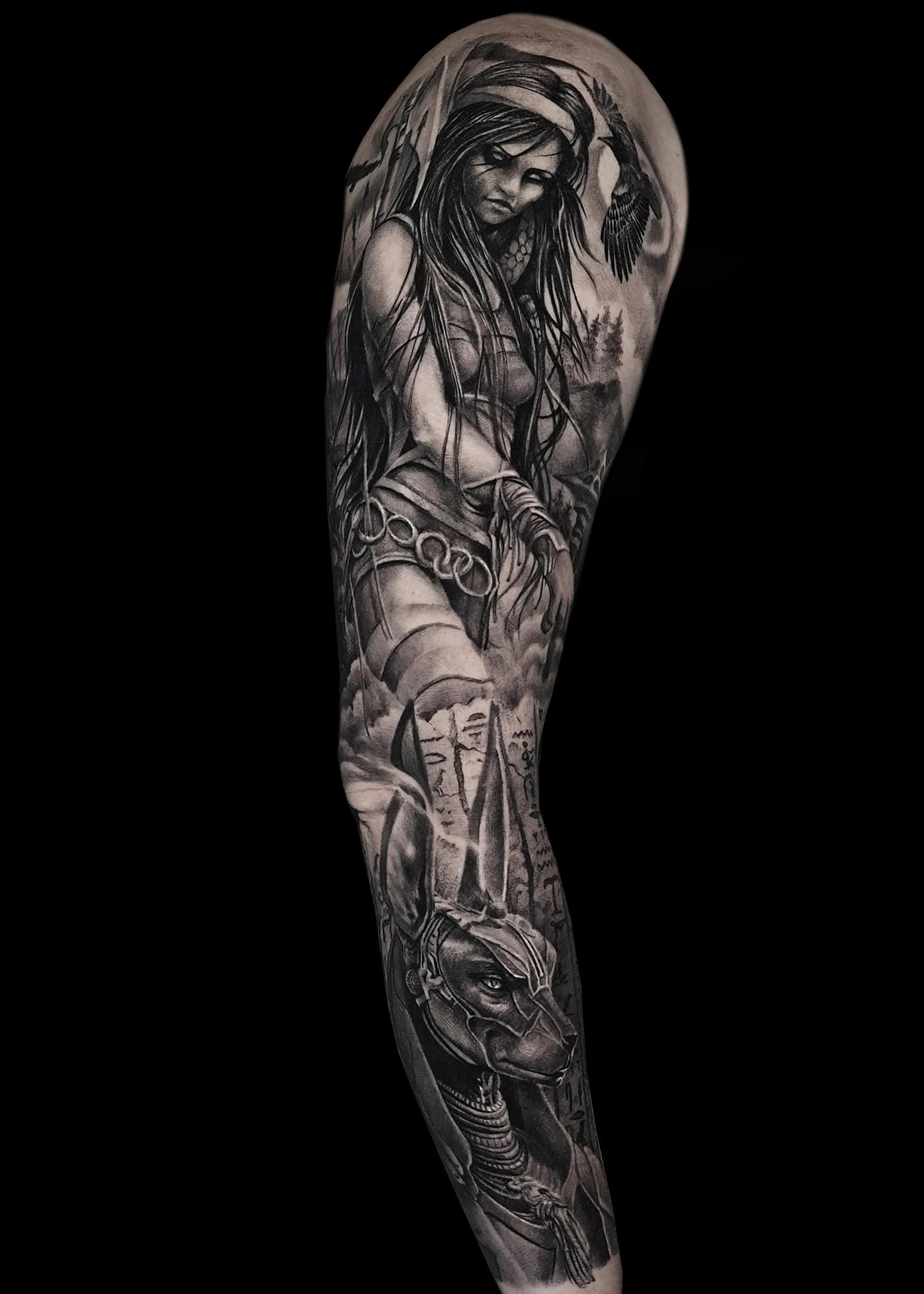 Czarno-szary rękaw tatuażu pokrywa ramię, przedstawiając szczegółową kobietę ozdobioną piórami we włosach i misternymi dodatkami. Poniżej przedstawiono rogatego, humanoidalnego człowieka z głową przypominającą zwierzę, na tle krajobrazu z drzewami, górami i zachmurzonym niebem.