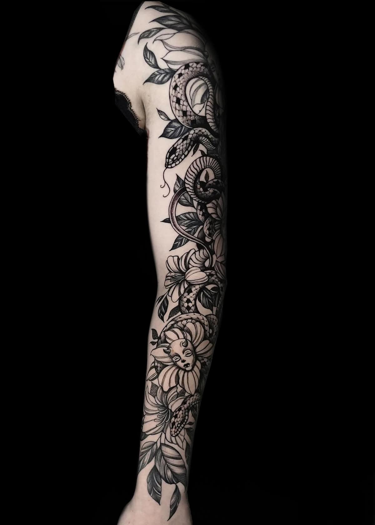 Przedramię ozdobione misternym czarno-szarym tatuażem na rękawie przedstawiającym zwiniętego węża, szczegółowe róże i różne liście. Grafika jest wyróżniona na jednolitym czarnym tle, podkreślając szczegółową pracę linii i cieniowanie wzoru tatuażu.