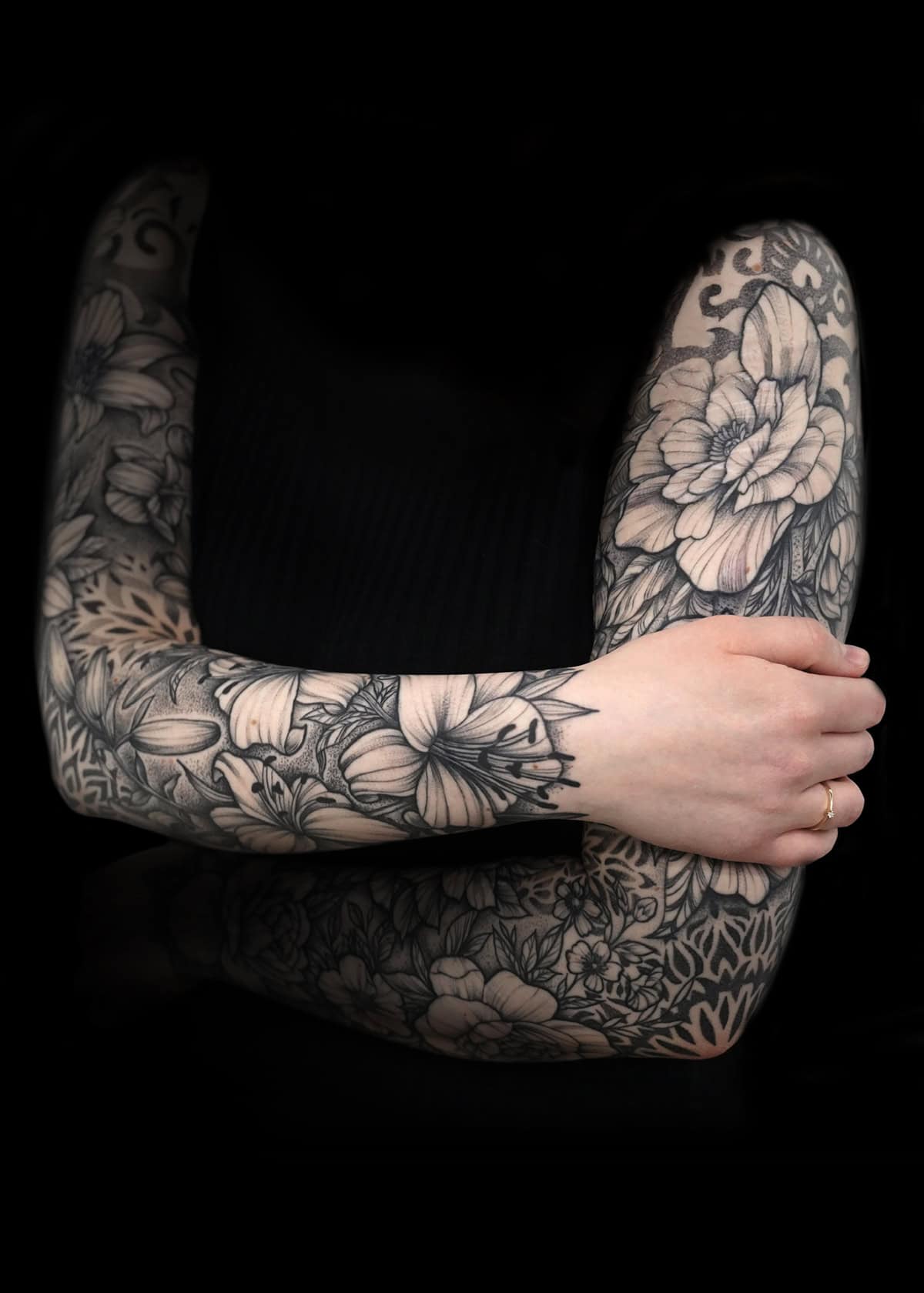 Osoba krzyżuje ramiona, prezentując szczegółowe czarno-białe tatuaże kwiatowe na ramionach na ciemnym tle. Tatuaże przedstawiają duże kwiaty, liście i skomplikowane wzory. Osoba nosi czarny top bez rękawów i złoty pierścień na palcu.
