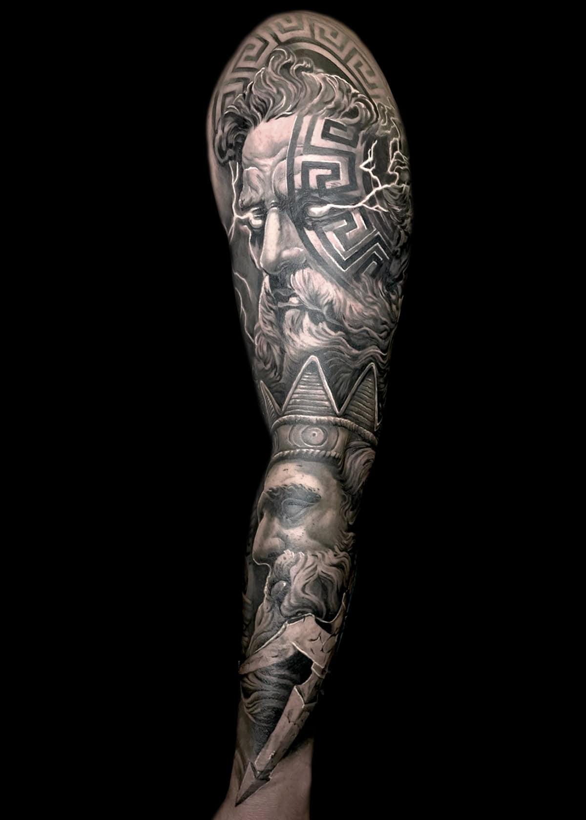 Szczegółowy czarno-szary tatuaż pokrywa całe ramię, przedstawiający klasyczny wizerunek twarzy brodatego mężczyzny z misternymi wzorami nad głową. Poniżej niego znajdują się różne inne elementy, w tym inna twarz i ręka ściskająca piorun, wszystkie wykonane z wysokim kontrastem i drobnymi szczegółami.
