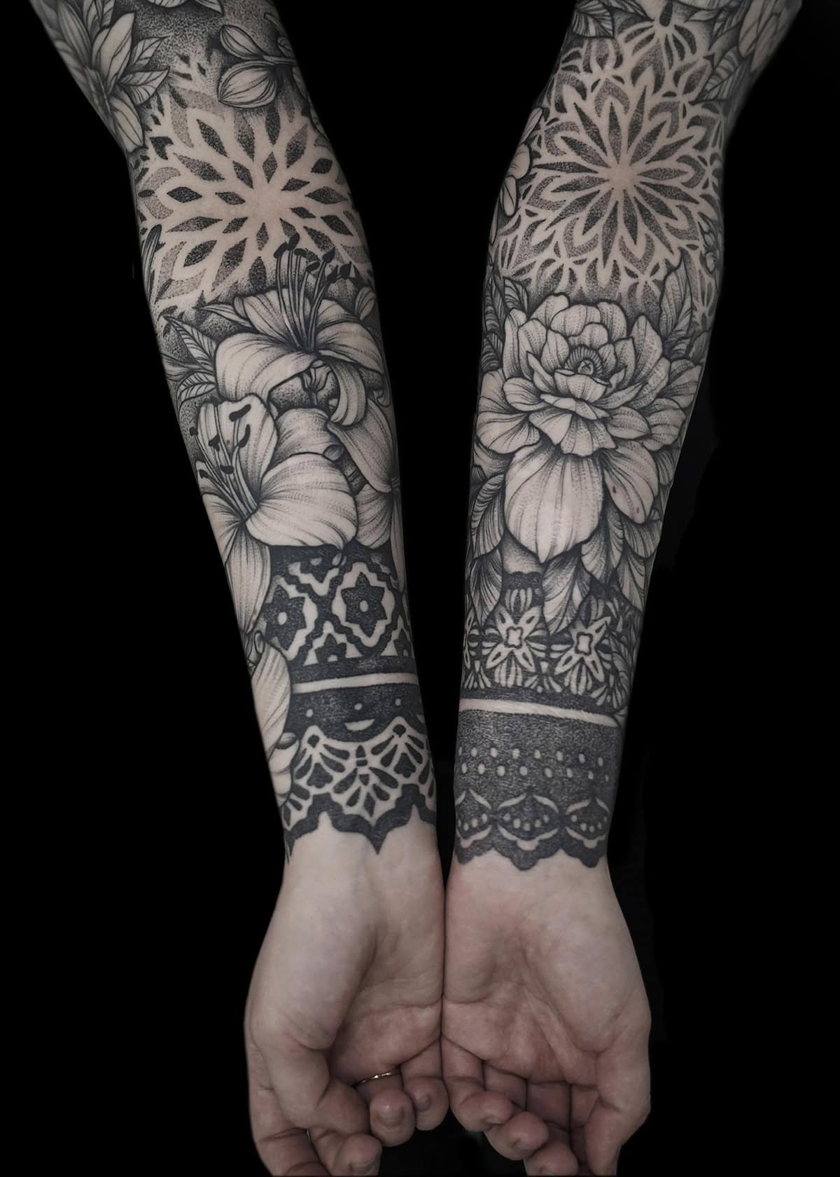 Na zdjęciu widać dwa mocno wytatuowane przedramiona z motywami kwiatowymi, w tym różami i liliami. Tatuaże są wykonane czarnym i szarym tuszem i rozciągają się od nadgarstków do łokci. Nadgarstki mają skomplikowane wzory przypominające koronkę, a przedramiona mają szczegółowe, duże główki kwiatów.