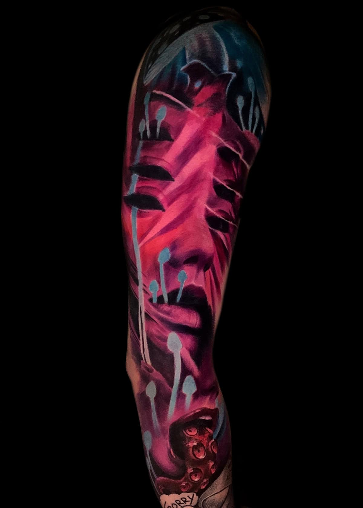 Żywy i surrealistyczny rękaw tatuażu pokrywa ramię z abstrakcyjną ludzką twarzą przedstawioną w odcieniach różu i fioletu. Niebieskie grzyby z wydłużonymi trzonami otaczają twarz, dodając kapryśnego kontrastu. Głębokie cienie i jasne refleksy podkreślają głębię i wymiar dzieła sztuki.